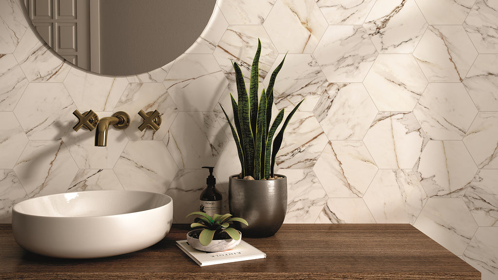 A white round vessel sink with gold fixtures and potted plants on a wooden counter against a white marble hexagon-tiled wall.