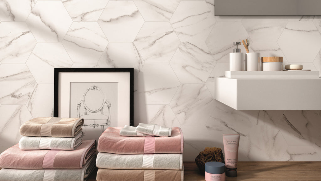 Stacks of towels and toiletries on a wooden counter next to a framed picture against a white marble hexagon-tiled wall.