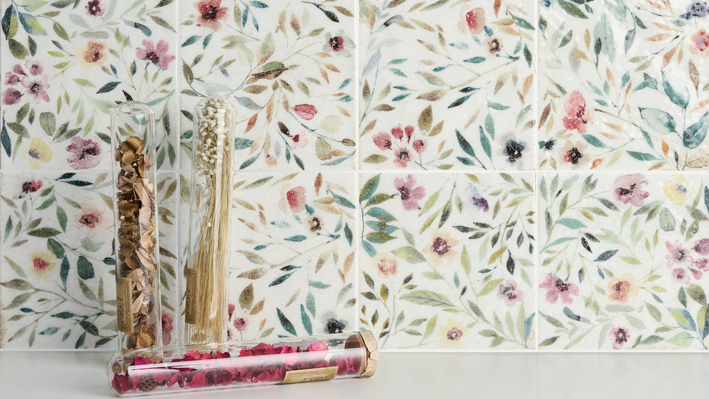 Glass tubes filled with dried botanicals stand on a white surface against a backdrop of watercolor-style floral patterned tiles.