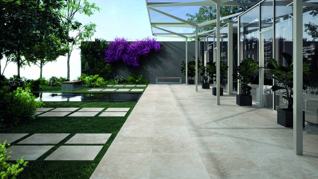 A wide outdoor courtyard view showing light beige stone pavers alongside a grassy lawn with a small water feature and glass storefronts.