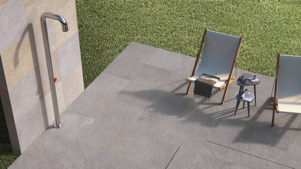 An elevated view of a gray tiled outdoor patio featuring a modern chrome shower, lounge chairs, and a small side table next to a grassy lawn.