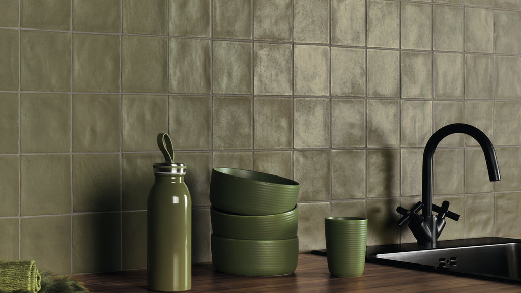 Green tableware and a water bottle sit on a dark wooden countertop next to a black sink and faucet, against an olive green square-tiled backsplash.