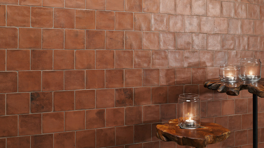 Lit candles in glass jars sit on rustic live-edge wood stands against a terracotta square-tiled wall.