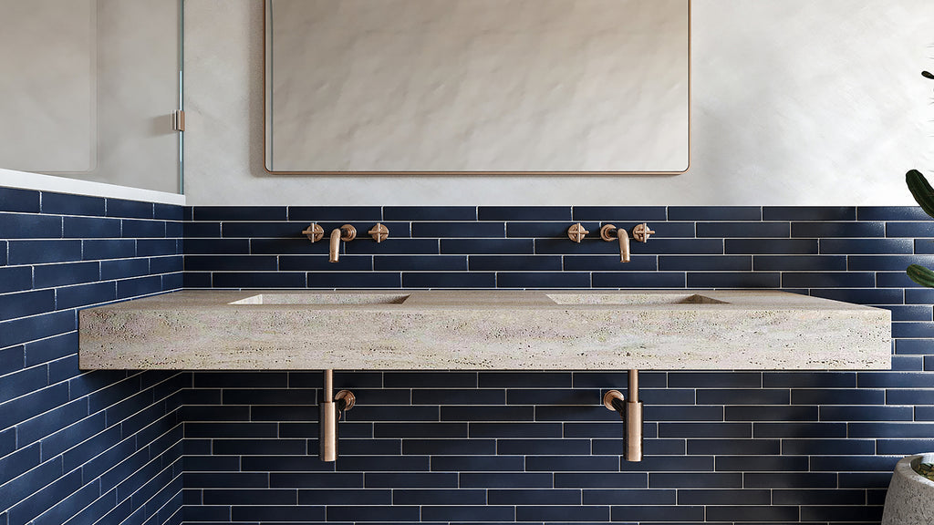 A double-sink stone vanity with copper fixtures mounted against a dark blue subway-tiled wall beneath a rectangular mirror.