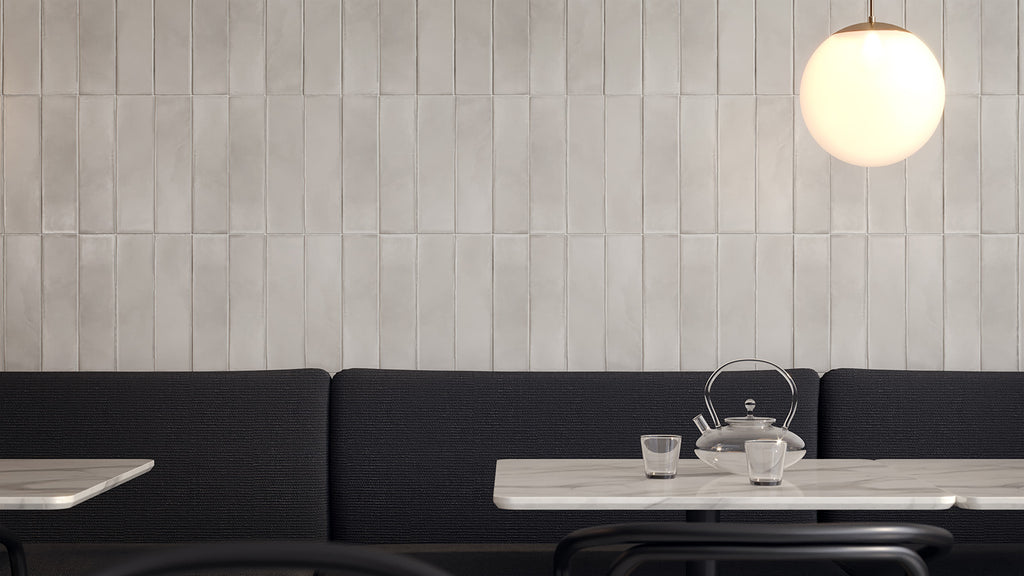 A glass teapot and cups sit on a marble table in front of a dark gray banquette and a light beige tiled wall with a glowing globe pendant light.