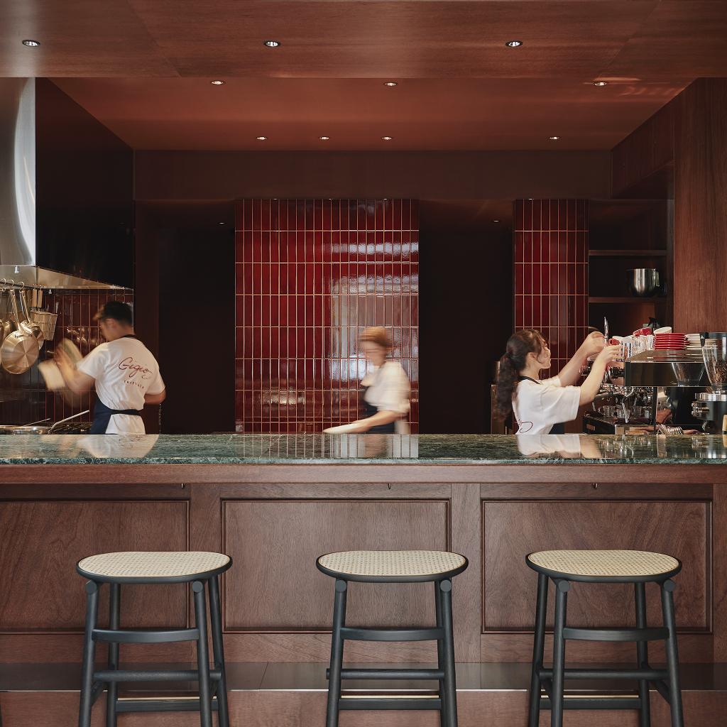 A bar-side view of a restaurant counter with black stools, featuring kitchen staff working behind it against a glossy red vertically tiled wall.
